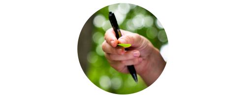 A hand holds a pen and leaf, the background of trees and green leaves is blurred. 